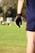 A person wearing a dark sports glove and shorts stands in a grassy field, with several people visible in the blurred background. The setting appears to be an outdoor sports field with a sunny atmosphere.