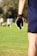 A person wearing a dark sports glove and shorts stands in a grassy field, with several people visible in the blurred background. The setting appears to be an outdoor sports field with a sunny atmosphere.