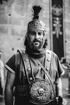 A historical figure dressed in ancient Roman attire, featuring a detailed helmet with embossed designs and a face guard. The individual has a serious expression and wears a tunic with decorative metalwork on the chest. The background includes a blurred banner with a design visible.