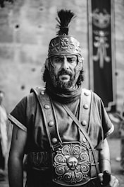 A historical figure dressed in ancient Roman attire, featuring a detailed helmet with embossed designs and a face guard. The individual has a serious expression and wears a tunic with decorative metalwork on the chest. The background includes a blurred banner with a design visible.