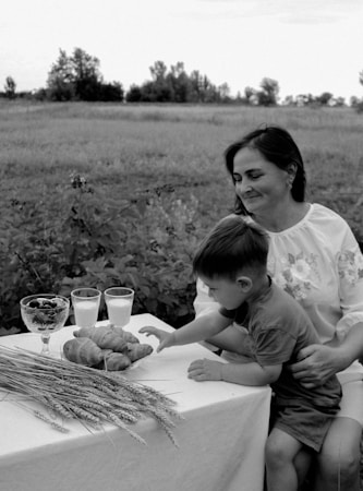 A black and white photograph depicts a woman and a child sitting at an outdoor table. The table is adorned with croissants, a bowl of fruits, two glasses of milk, and some wheat stalks. The background shows an open field with trees in the distance, providing a serene and pastoral setting.