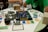 A tabletop board game setup is laid out on a circular table with a white cloth cover. The game involves a grid board with the word 'ACQUIRE' at the bottom, featuring various plastic game pieces shaped like buildings, and several cards organized around the board. Two people are partially visible, one in a green shirt and the other in a striped shirt, possibly engaged in playing the game. Several glasses, one containing a dark liquid, are on the table alongside paper currency and game cards of different colors.