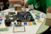 A tabletop board game setup is laid out on a circular table with a white cloth cover. The game involves a grid board with the word 'ACQUIRE' at the bottom, featuring various plastic game pieces shaped like buildings, and several cards organized around the board. Two people are partially visible, one in a green shirt and the other in a striped shirt, possibly engaged in playing the game. Several glasses, one containing a dark liquid, are on the table alongside paper currency and game cards of different colors.
