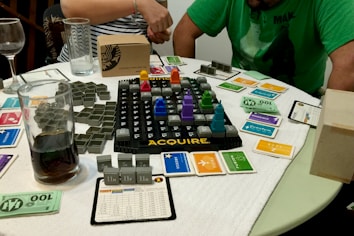 A tabletop board game setup is laid out on a circular table with a white cloth cover. The game involves a grid board with the word 'ACQUIRE' at the bottom, featuring various plastic game pieces shaped like buildings, and several cards organized around the board. Two people are partially visible, one in a green shirt and the other in a striped shirt, possibly engaged in playing the game. Several glasses, one containing a dark liquid, are on the table alongside paper currency and game cards of different colors.