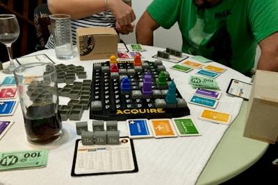 A tabletop board game setup is laid out on a circular table with a white cloth cover. The game involves a grid board with the word 'ACQUIRE' at the bottom, featuring various plastic game pieces shaped like buildings, and several cards organized around the board. Two people are partially visible, one in a green shirt and the other in a striped shirt, possibly engaged in playing the game. Several glasses, one containing a dark liquid, are on the table alongside paper currency and game cards of different colors.