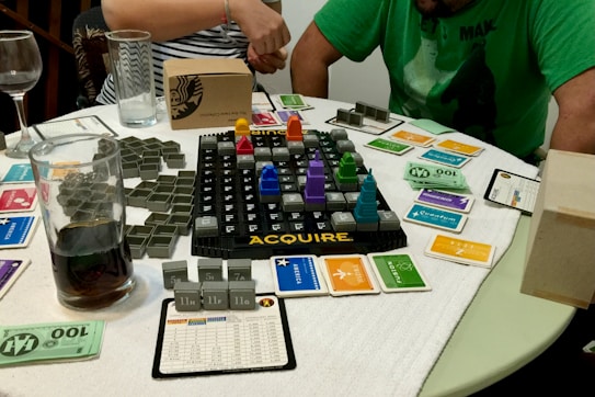 A tabletop board game setup is laid out on a circular table with a white cloth cover. The game involves a grid board with the word 'ACQUIRE' at the bottom, featuring various plastic game pieces shaped like buildings, and several cards organized around the board. Two people are partially visible, one in a green shirt and the other in a striped shirt, possibly engaged in playing the game. Several glasses, one containing a dark liquid, are on the table alongside paper currency and game cards of different colors.
