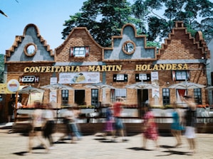 A building with Dutch architectural style, featuring decorative gables and brickwork, serves as a pastry shop with signs in Portuguese. People are walking by in a blur, indicating motion, and there are several umbrellas outside the establishment.