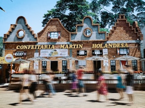 A building with Dutch architectural style, featuring decorative gables and brickwork, serves as a pastry shop with signs in Portuguese. People are walking by in a blur, indicating motion, and there are several umbrellas outside the establishment.