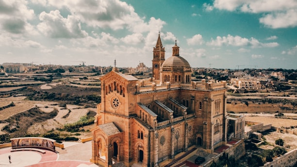 A magnificent church with intricate architecture stands prominently in a rural landscape. The structure features a large dome and two tall towers with crosses. Surrounding the church are expansive fields and distant buildings under a partly cloudy sky.