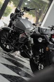 A shiny motorcycle is displayed in a showroom with a sleek, modern design. The motorcycle has a matte finish and detailed mechanical components. It is parked on a checkered floor, and there's a plastic cover over the seat indicating it's brand new. Large windows in the background allow natural light to illuminate the scene.