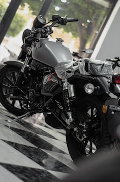 A shiny motorcycle is displayed in a showroom with a sleek, modern design. The motorcycle has a matte finish and detailed mechanical components. It is parked on a checkered floor, and there's a plastic cover over the seat indicating it's brand new. Large windows in the background allow natural light to illuminate the scene.