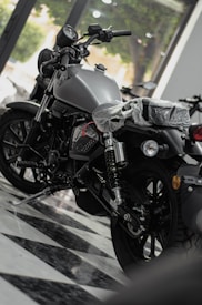 A shiny motorcycle is displayed in a showroom with a sleek, modern design. The motorcycle has a matte finish and detailed mechanical components. It is parked on a checkered floor, and there's a plastic cover over the seat indicating it's brand new. Large windows in the background allow natural light to illuminate the scene.