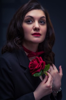 A woman with long, dark hair holds a red rose near her chest, wearing a black outfit with a red neck accessory. Her expression is serious and contemplative.