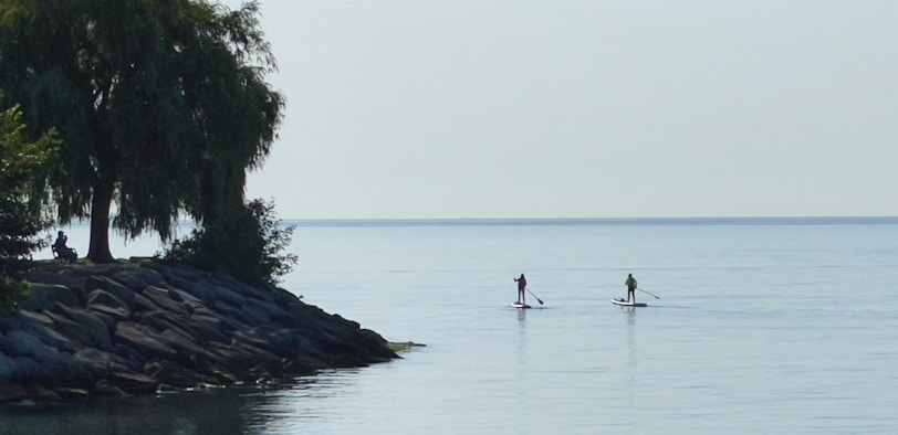 Two people paddling a sleek electric stand up paddleboard catamaran on calm blue water under a clear sky.