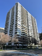 A tall, modern apartment building with multiple balconies and abundant greenery adorning the facade. The structure is white with large, rectangular windows and is set against a clear, blue sky. Sparse trees with bare branches are visible at the street level, and a few cars can be seen parked along the road.