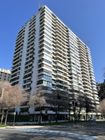 A tall, modern apartment building with multiple balconies and abundant greenery adorning the facade. The structure is white with large, rectangular windows and is set against a clear, blue sky. Sparse trees with bare branches are visible at the street level, and a few cars can be seen parked along the road.
