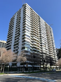 A tall, modern apartment building with multiple balconies and abundant greenery adorning the facade. The structure is white with large, rectangular windows and is set against a clear, blue sky. Sparse trees with bare branches are visible at the street level, and a few cars can be seen parked along the road.