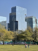 A modern cityscape featuring tall, reflective glass buildings against a clear blue sky. In the foreground, a grassy park area with several people enjoying outdoor activities. Trees with green foliage partially obscure the buildings.