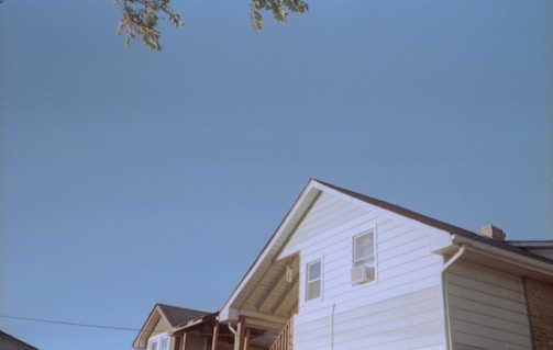 A low-angle view of the upper part of a house with white siding and a pitched roof. The clear blue sky occupies most of the frame, with a branch with a few leaves extending from the top left corner.