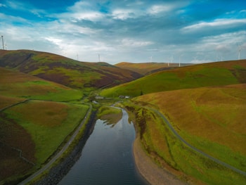 A serene landscape featuring rolling green and brown hills under a partly cloudy sky. Several wind turbines are scattered across the hills in the background. A calm river flows through the center of the scene, with a small road running alongside it.