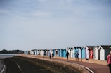 A row of colorful beach huts lines a seaside promenade with a few people walking along the path. The sky is clear and the sea is calm, enhancing the serene coastal atmosphere.