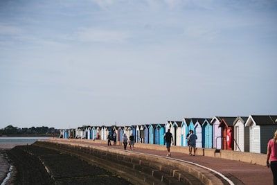 A row of colorful beach huts lines a seaside promenade with a few people walking along the path. The sky is clear and the sea is calm, enhancing the serene coastal atmosphere.