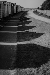 A row of small cabins or beach huts with pitched roofs lines a paved path. The path is bordered by grass and a gravel walkway, with shadows cast across both surfaces. Sparse vegetation is visible on the right side of the image.