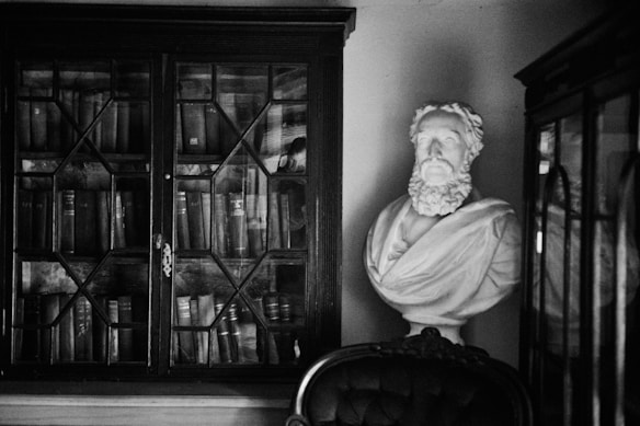A black and white photograph captures a classic, scholarly ambiance with a bust of a bearded man on a pedestal beside a wooden cabinet filled with books. The glass-fronted cabinet shows the spines of various thick volumes. An old, upholstered chair partially appears in the foreground, adding to the vintage feel.