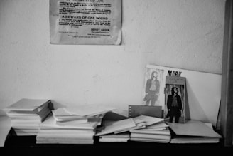 A black and white scene with a stack of papers on a flat surface and several photographs propped against a wall. The photographs include images of a person in a suit. There is a notice or poster pinned to the wall above.