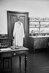A black and white photograph depicting an old-fashioned apothecary or pharmacy room. A white lab coat hangs on a wooden door. On one side, shelves with jars and bottles, potentially containing various substances, line the wall. A wooden desk and chair are situated in the foreground, with various items such as a bottle, an open book, and small containers placed on it. The room has a rustic look with wooden furniture and fixtures.