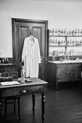 A black and white photograph depicting an old-fashioned apothecary or pharmacy room. A white lab coat hangs on a wooden door. On one side, shelves with jars and bottles, potentially containing various substances, line the wall. A wooden desk and chair are situated in the foreground, with various items such as a bottle, an open book, and small containers placed on it. The room has a rustic look with wooden furniture and fixtures.