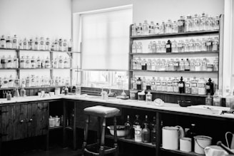 A vintage apothecary or laboratory setting showcasing wooden shelves lined with numerous glass bottles and jars. The bottles have labels and are neatly arranged on dark wooden shelves. A wooden counter runs along the walls with various laboratory equipment, including flasks and beakers. A high stool is placed in front of the counter, and a window with a shade partially drawn allows natural light to enter.
