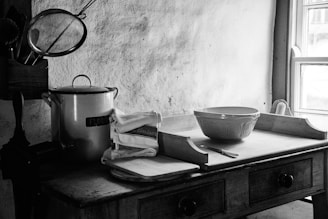 A vintage kitchen setup with a wooden table, holding a large ceramic mixing bowl, a metal container labeled 'Flour', and some folded dish towels. There is a window on the right side allowing natural light in, casting soft shadows. A strainer and cooking utensils are placed on the side, and the overall setting suggests a rustic, old-fashioned kitchen.