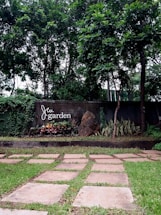 A lush garden featuring a well-maintained pathway made of flat stones on a grassy lawn. The background is dominated by tall trees and a concrete wall with the words 'Jai garden' displayed on it. There is also a rocky feature with various plants and shrubs around it.