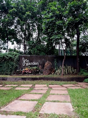 A lush garden featuring a well-maintained pathway made of flat stones on a grassy lawn. The background is dominated by tall trees and a concrete wall with the words 'Jai garden' displayed on it. There is also a rocky feature with various plants and shrubs around it.