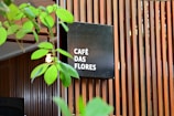 A modern wooden slatted wall serves as a backdrop for a black square sign displaying the words 'CAFÉ DAS FLORES' in white text. Green plant leaves partially obscure the view, adding a natural element to the scene. The overall setting gives the impression of an inviting, stylish café atmosphere.