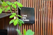 A modern wooden slatted wall serves as a backdrop for a black square sign displaying the words 'CAFÉ DAS FLORES' in white text. Green plant leaves partially obscure the view, adding a natural element to the scene. The overall setting gives the impression of an inviting, stylish café atmosphere.