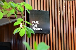 A modern wooden slatted wall serves as a backdrop for a black square sign displaying the words 'CAFÉ DAS FLORES' in white text. Green plant leaves partially obscure the view, adding a natural element to the scene. The overall setting gives the impression of an inviting, stylish café atmosphere.