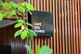 A modern wooden slatted wall serves as a backdrop for a black square sign displaying the words 'CAF&Eacute; DAS FLORES' in white text. Green plant leaves partially obscure the view, adding a natural element to the scene. The overall setting gives the impression of an inviting, stylish caf&eacute; atmosphere.