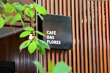 A modern wooden slatted wall serves as a backdrop for a black square sign displaying the words 'CAFÉ DAS FLORES' in white text. Green plant leaves partially obscure the view, adding a natural element to the scene. The overall setting gives the impression of an inviting, stylish café atmosphere.