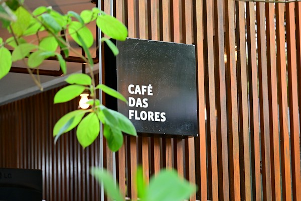 A modern wooden slatted wall serves as a backdrop for a black square sign displaying the words 'CAF&Eacute; DAS FLORES' in white text. Green plant leaves partially obscure the view, adding a natural element to the scene. The overall setting gives the impression of an inviting, stylish caf&eacute; atmosphere.