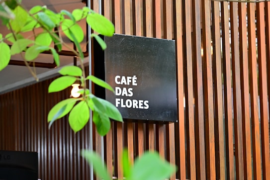 A modern wooden slatted wall serves as a backdrop for a black square sign displaying the words 'CAF&Eacute; DAS FLORES' in white text. Green plant leaves partially obscure the view, adding a natural element to the scene. The overall setting gives the impression of an inviting, stylish caf&eacute; atmosphere.