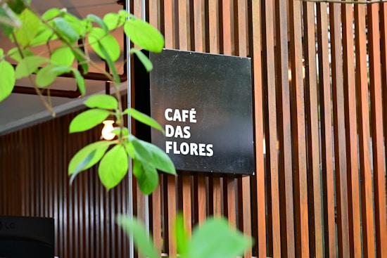 A modern wooden slatted wall serves as a backdrop for a black square sign displaying the words 'CAF&Eacute; DAS FLORES' in white text. Green plant leaves partially obscure the view, adding a natural element to the scene. The overall setting gives the impression of an inviting, stylish caf&eacute; atmosphere.
