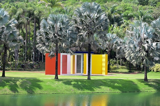 A modern, colorful structure with vibrant, vertical panels of red, white, yellow, and orange surrounded by tall palm trees. The lush greenery is reflected in a calm body of water in the foreground, creating a peaceful and harmonious atmosphere.