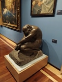 A museum exhibit featuring a sculpture of a seated, contemplative figure made of dark material on a pedestal. Behind the sculpture, there are two framed paintings hanging on a blue wall. The sculpture is positioned on a polished wooden floor.