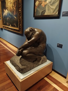 A museum exhibit featuring a sculpture of a seated, contemplative figure made of dark material on a pedestal. Behind the sculpture, there are two framed paintings hanging on a blue wall. The sculpture is positioned on a polished wooden floor.