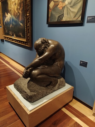 A museum exhibit featuring a sculpture of a seated, contemplative figure made of dark material on a pedestal. Behind the sculpture, there are two framed paintings hanging on a blue wall. The sculpture is positioned on a polished wooden floor.