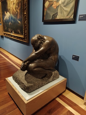 A museum exhibit featuring a sculpture of a seated, contemplative figure made of dark material on a pedestal. Behind the sculpture, there are two framed paintings hanging on a blue wall. The sculpture is positioned on a polished wooden floor.