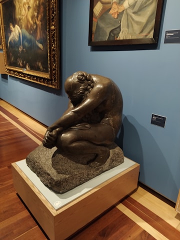 A museum exhibit featuring a sculpture of a seated, contemplative figure made of dark material on a pedestal. Behind the sculpture, there are two framed paintings hanging on a blue wall. The sculpture is positioned on a polished wooden floor.