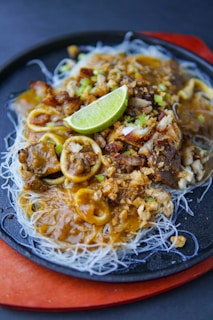 A dish of thin rice noodles topped with a rich brown sauce, garnished with fried garlic, crispy bits, and slices of lime sits on a dark round plate. Small pieces of meat and calamari rings are mixed in, adding texture and flavor to the dish.